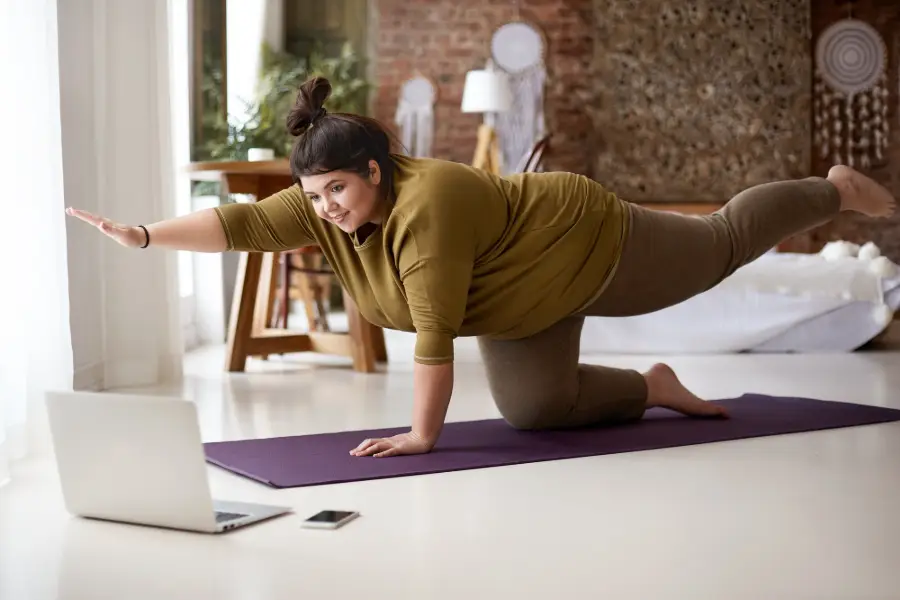Obese chubby young European female with hair knot practicing yoga or pilates indoors on mat, doing exercises to strengthen core, watching video lesson online in front of open laptop computer on floor